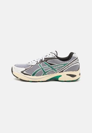 Zapatillas deportivas con parte superior de malla en blanco y negro, acentuadas por detalles en verde. Presentan una suela acolchada y un sistema de cordones visible.