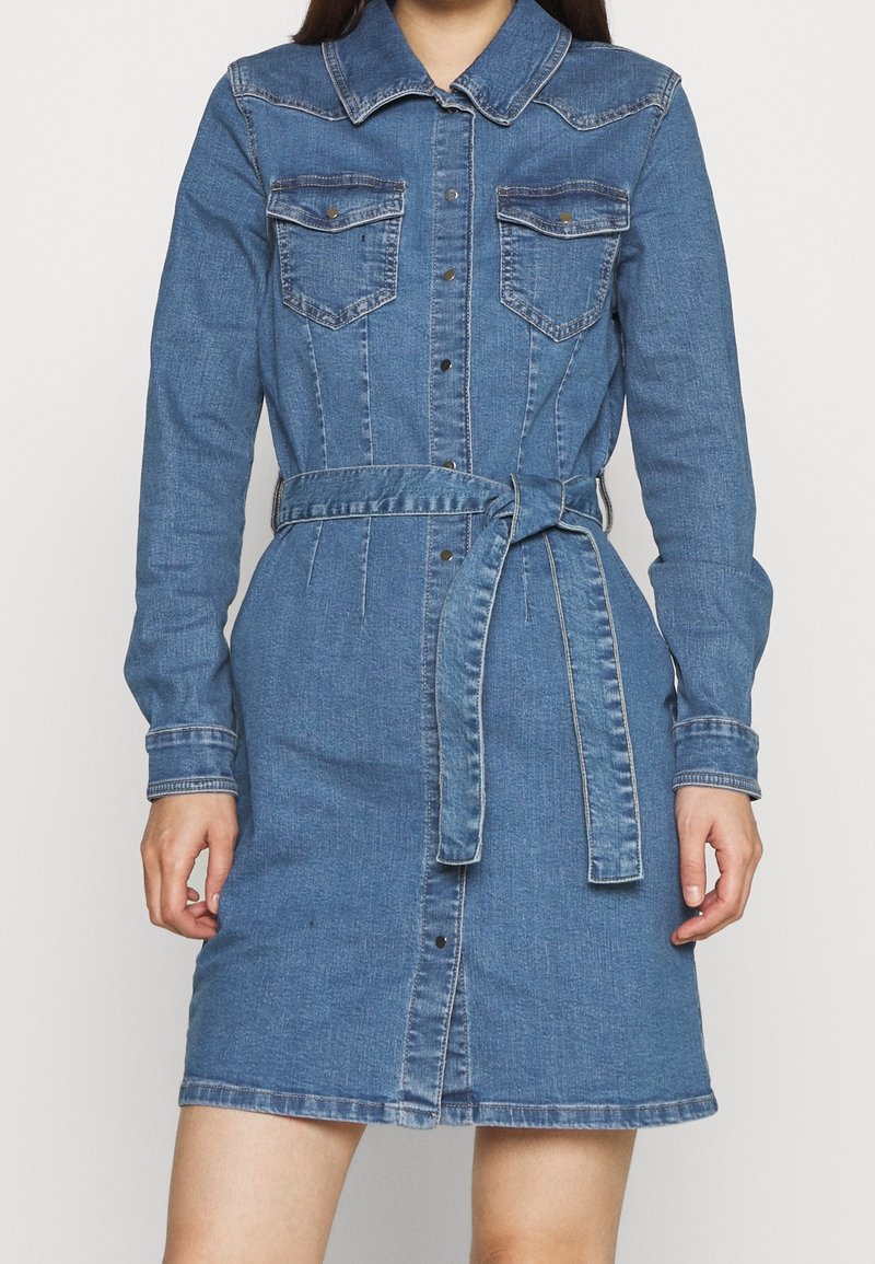 Femme portant une robe en denim bleu à manches longues avec des boutons sur le devant, deux poches poitrine et une ceinture nouée à la taille, debout devant un fond uni.