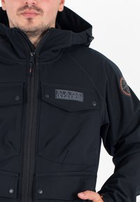 Man in een zwart jack met rits, met een 'NAPA' logo patch op de borstzak en een ronde patch op de bovenste mouw.