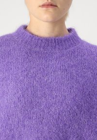 Närbild av en person som bär en mjuk, lila stickad tröja med ribbad rund halsringning, med fokus på bröst- och nackområdet.