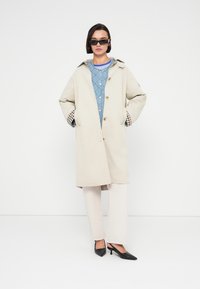 Μπεζ αδιάβροχο με γιακά, διακοσμημένο με καφέ κουμπιά και μανσέτες με καρώ σχέδιο, φορεμένο πάνω από μια μπλε κουμπωτή μπλούζα και ανοιχτόχρωμο παντελόνι.