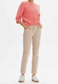 Jasnoróżowy dziergany sweter z teksturowanym wzorem, zestawiony z jasnobeżowymi spodniami ze sznurkiem w pasie. Całość dopełniają białe trampki.