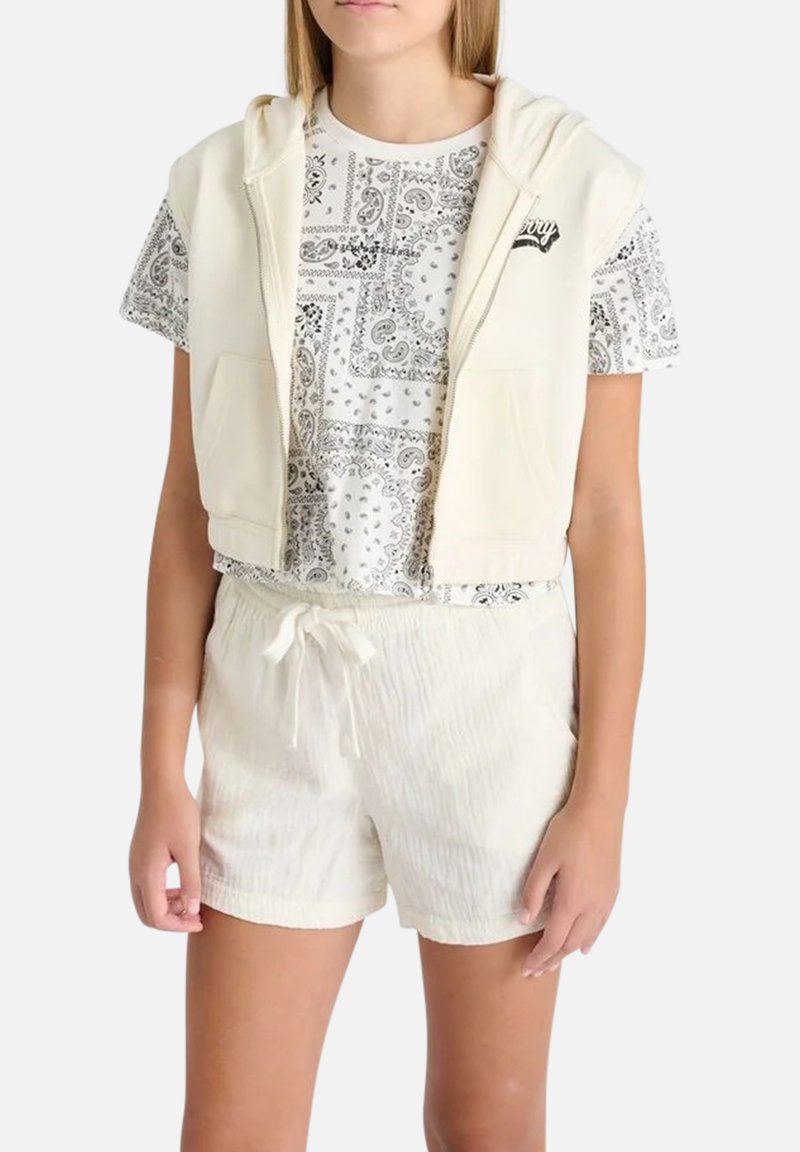 Hoodie crème coupé avec poches par-dessus une chemise à motif paisley noir et blanc, associé à un short léger texturé avec cordon de serrage.