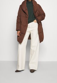 Hnědá péřová bunda s velkým límcem, spárovaná se tmavě zeleným svetrem a světlými béžovými širokými kalhotami. Jednoduchý design, texturovaná tkanina.