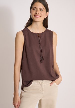 Femme aux longs cheveux bruns porte un haut marron sans manches avec des franges et un pantalon beige clair, debout et souriante avec une main dans la poche.