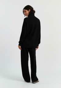 Zwarte trui met een hoge kraag en geribbelde textuur, gecombineerd met loszittende zwarte broeken, wat een samenhangende monochrome uitstraling creëert.