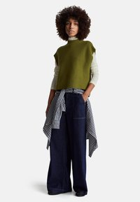 Žena s kudrnatými vlasy, oblečená v olivově zeleném svetru bez rukávů, bílém krajkovém tričku s dlouhým rukávem, širokých džínech a s kostičkovanou košilí uvázanou v pase.