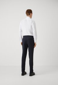 Camicia bianca a maniche lunghe abbinata a pantaloni scuri blu navy, caratterizzati da una texture liscia e un design aderente. Scarpe nere completano l'outfit.