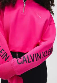Svetle růžová zipová mikina vyrobená z měkké tkaniny. Obsahuje žebrované manžety s nápisem "CALVIN KLEIN" vytištěným černou barvou na rukávech.