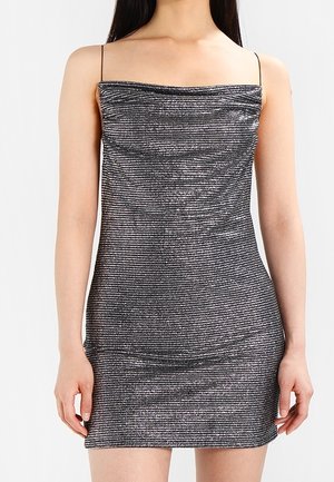 Femme portant une mini robe argentée métallique sans manches avec de fines bretelles spaghetti et un décolleté légèrement drapé.