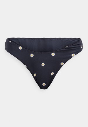 Fekete bikini alsó fehér és sárga százszorszép mintázattal és hullámmintás díszítéssel a derékvonalon, síkban elhelyezve egy fehér háttér előtt.