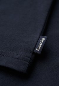 Detalles de una camiseta de algodón color navy con mangas de doble costura y una etiqueta de marca en texto blanco sobre un fondo navy.