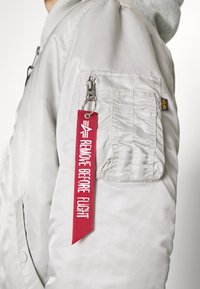Svetlošedá bomber bunda, nylonový materiál, rebrovaný golier, zips na vrecku na ľavom rukáve, obsahuje červenú visačku s textom "ODSTRÁNIŤ PRED LETOM."