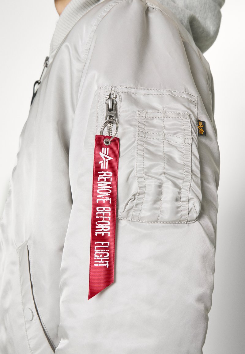 Veste bomber gris clair en nylon, col côtelé, poche zippée sur la manche gauche, avec une étiquette rouge portant le texte "ENLEVER AVANT LE VOL."