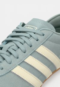 Ανοιχτά μπλε σουέτ αθλητικά παπούτσια με λευκές ζιγκ-ζαγκ ρίγες, στρογγυλά κορδόνια και ανάγλυφο λογότυπο στη γλώσσα. Λαστιχένια σόλα.