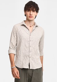 Camicia button-up beige chiaro con motivo geometrico testurizzato, maniche arrotolate e colletto classico. Bottoni in tonalità argento e una leggera lucentezza.
