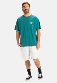 Camiseta de algodón color verde azulado con mangas cortas y cuello redondo, que presenta un diseño gráfico en el pecho. Combinada con pantalones cortos blancos y zapatillas negras.