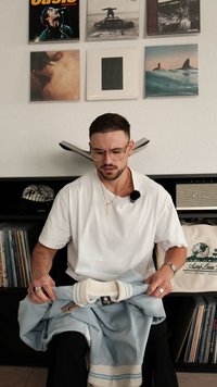 Ljusblå långärmad skjorta med vita ränder på kragen och ärmsluten, hålls av en person som bär en vit t-shirt; vinylskivor och högtalare i bakgrunden.