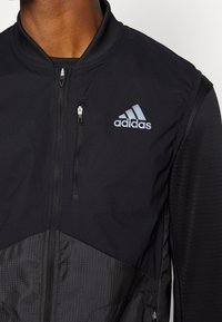 Black Adidas jacket with a zippered front, featuring a smooth upper fabric and textured lower section with a grid pattern. Logo on chest.