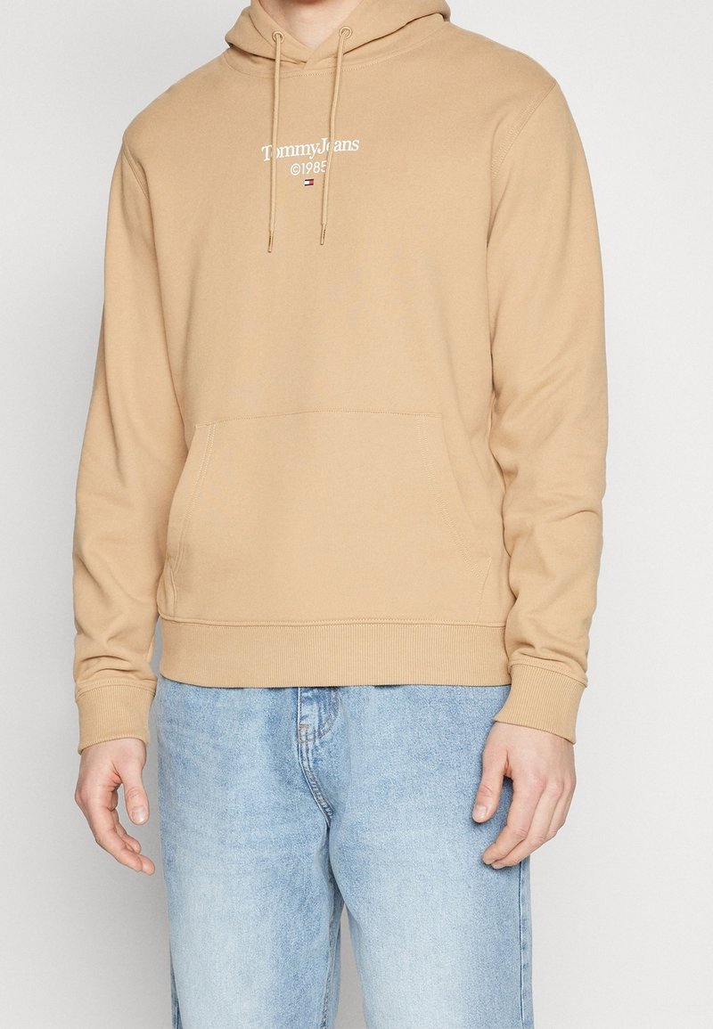Homme portant un sweat à capuche beige Tommy Jeans avec poche avant et jean bleu clair, debout devant un fond clair uni.