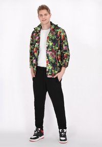 Jachetă multicoloră cu model de frunze tropicale, pantaloni negri și adidași înalți. Jacheta are glugă și manșete elastice.