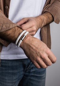 Balts nopīts ādas aproce ar metāla sprādzi, uzlikta uz rokas, izceļot teksturētu dizainu uz neitrāla fona.