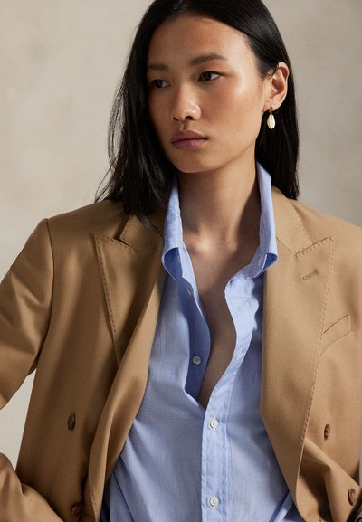 Kamelový blejzer s ostrými klopami over light blue button-up košele. Blejzer má hladkú textúru a dvojradové zapínanie; jemné detaily stehov sú viditeľné.