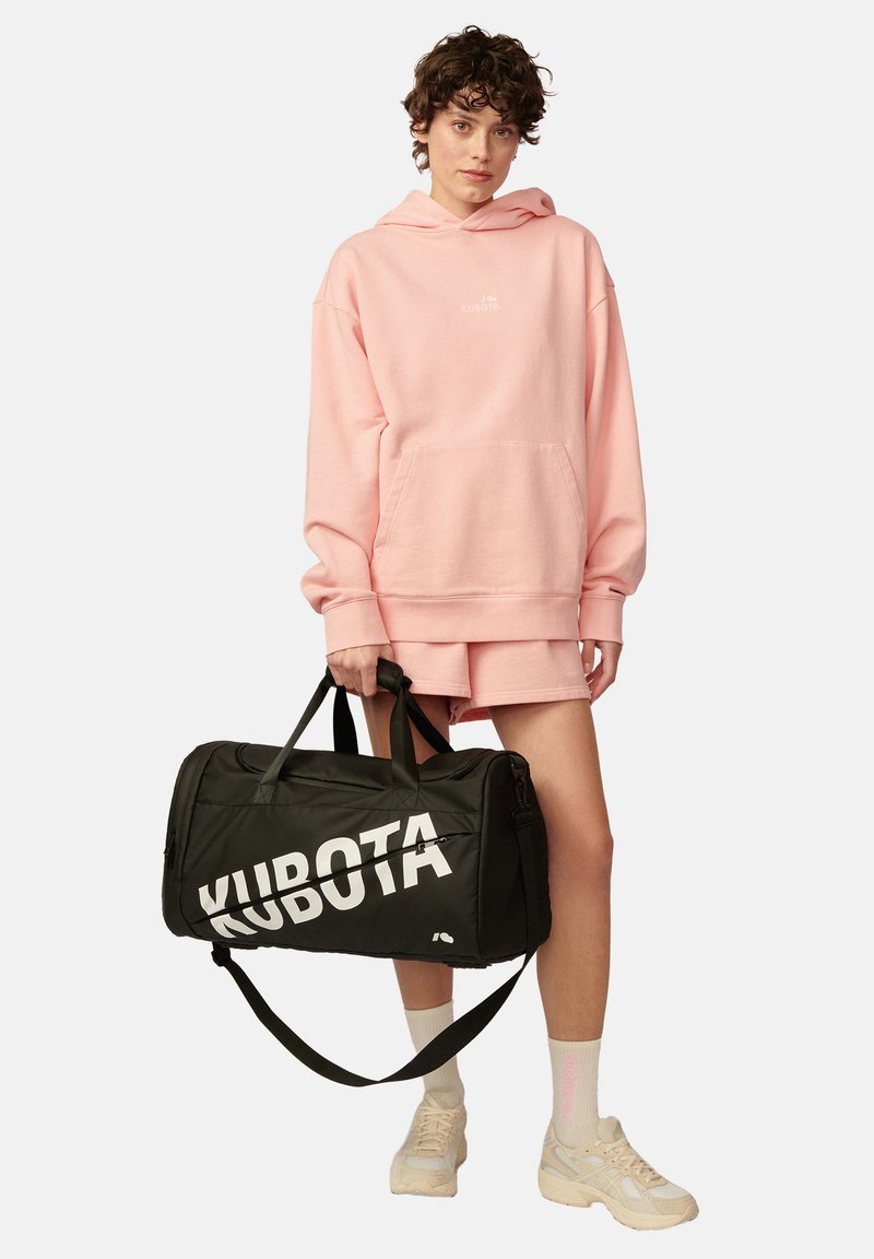 Różowa bluza z kapturem i kieszenią z przodu, z białym logo, zestawiona z czarną torbą podróżną z wyraźnym białym napisem, noszona z jasnymi trampkami i pastelowymi skarpetkami.