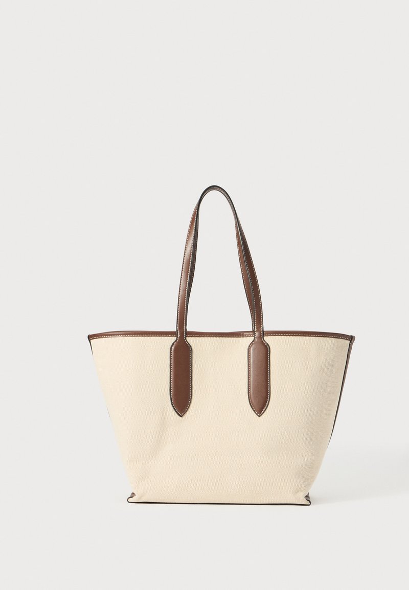 Beige canvas tote bag with brown leather handles and trim. Features a wide, open top and a structured, tapered shape. White stitching accents.
