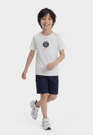 Garçon souriant marchant, portant un t-shirt blanc du Paris Saint-Germain, un short cargo marine et des baskets blanches New Balance, sur un fond uni.