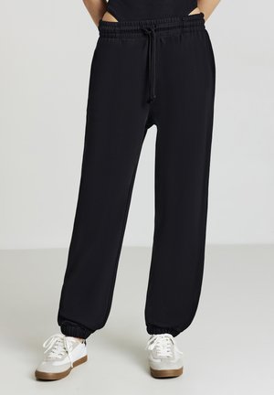 Pantalons de jogging noirs avec une taille à cordon, des poignets élastiques et des poches latérales. Associés à des baskets de couleur claire sur un fond neutre.