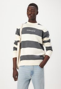 Csíkos pulóver szürke és krém színben, kötött anyagból, kerek nyakkivágással, bordázott mandzsettákkal és aljjal. A mellkason található a "GUESS JEANS" logó.