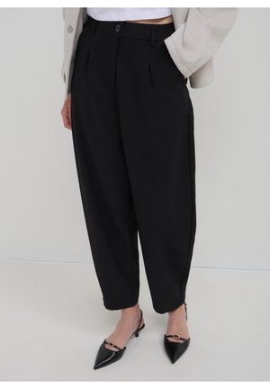 Mujer con pantalones negros holgados, cárdigan beige, top blanco corto y tacones negros de punta con tira trasera, de pie frente a un fondo liso.