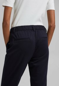Personne portant un pantalon noir à taille élastique avec des poches arrière et une chemise blanche à manches courtes rentrée, debout devant un fond uni.