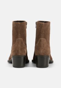Botas de ante marrón hasta el tobillo con un tacón estrecho, con un cierre de cremallera en el lateral y sutiles detalles de costura. Suelas negras lisas.
