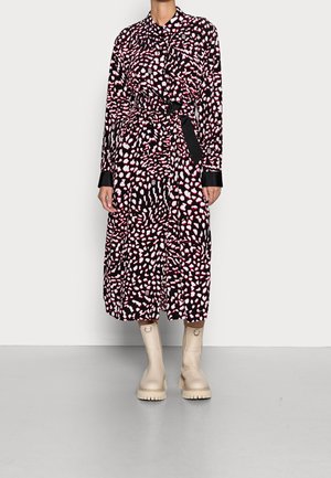Mujer que lleva un vestido negro de manga larga con manchas abstractas blancas y rojas, cintura ceñida con cinturón y botas beige gruesas hasta la mitad de la pantorrilla, de pie sobre un suelo gris.