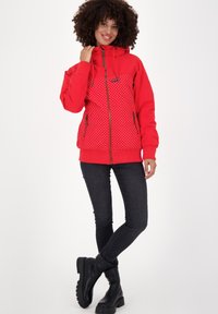 Glunderende vrouw met krullend haar, gekleed in een rode hoodie-jack met witte stippen, zwarte skinny jeans en zwarte laarzen, staande met gekruiste benen.