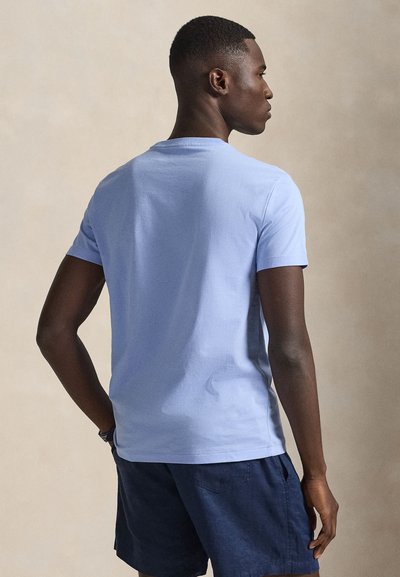 Ανοιχτό μπλε κοντομάνικο βαμβακερό μπλουζάκι με στρογγυλή λαιμόκοψη και άνετη εφαρμογή, εμφανιζόμενο από πίσω με ουδέτερο φόντο.