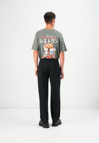 Graues T-Shirt mit Cartoon-Hund-Grafik und japanischem Text auf dem Rücken; kombiniert mit schwarzen Plisseehosen und klobigen schwarzen Schuhen.