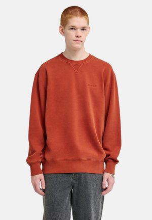 Jonge man met kort rood haar die een verbrande oranje trui en grijze spijkerbroek draagt, staand tegen een effen lichte achtergrond.