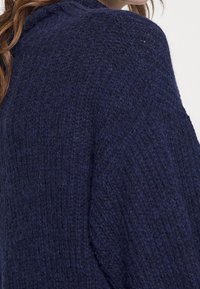 Marinblå stickad tröja med ett texturerat mönster, som har en avslappnad passform och nedbundna axelsömmar. Mjuk, fluffig garn synlig i väven.