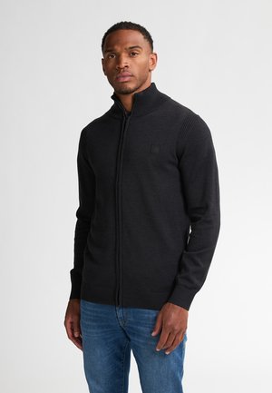Man in een zwarte rits trui met geribbelde kraag en mouwen, gecombineerd met een blauwe jeans, staand tegen een effen lichte achtergrond.