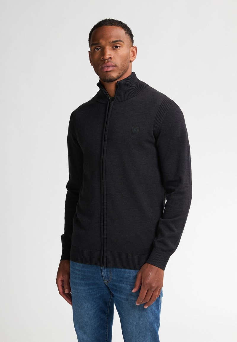 Mann in einem schwarzen Reißverschluss-Pullover mit geripptem Kragen und Bündchen, kombiniert mit blauen Jeans, steht vor einem schlichten hellen Hintergrund.