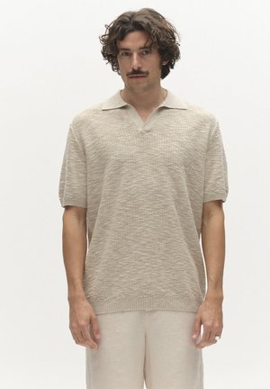 Muž kučeravých vlasov a s fúzmi, oblečený v béžovej krátkorukávovej textúrovanej pletenej polokošeli a svetlých nohaviciach, stojaci na bielom pozadí.