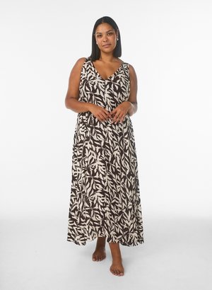 Frau steht barfuß in einem ärmellosen, schwarz-weißen Maxikleid mit Blattmuster vor einem schlichten weißen Hintergrund.