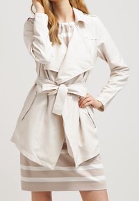 Jasno beżowy płaszcz trench z paskiem, z oversize'owymi klapami i kieszeniami po bokach, nałożony na sukienkę w paski w neutralnych odcieniach.