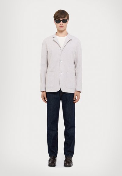 Helles graues Strickblazer mit einem einzigen Knopf, Fronttaschen, kombiniert mit dunkelblauen Jeans und einer schwarzen Sonnenbrille.