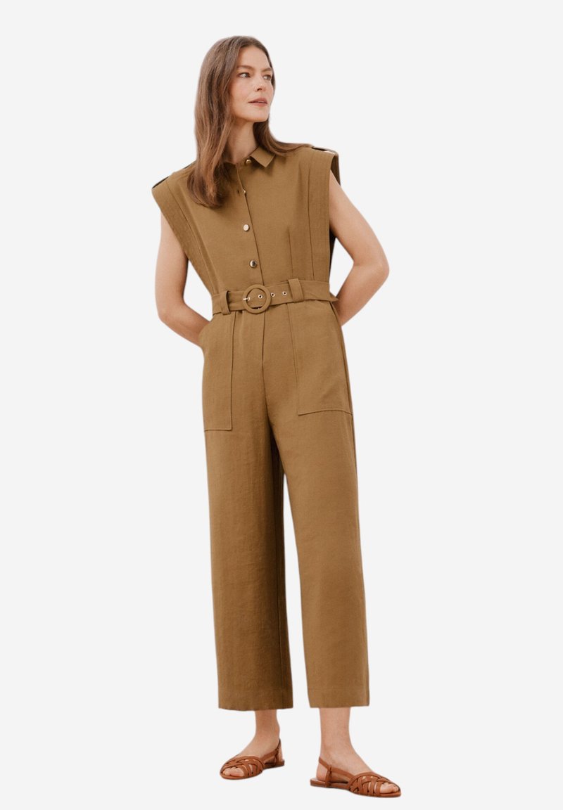 Kaki jumpsuit med axelepåler, knappad front, bälte i midjan, vida ben och stora fickor. Kombinerad med bruna platta sandaler.
