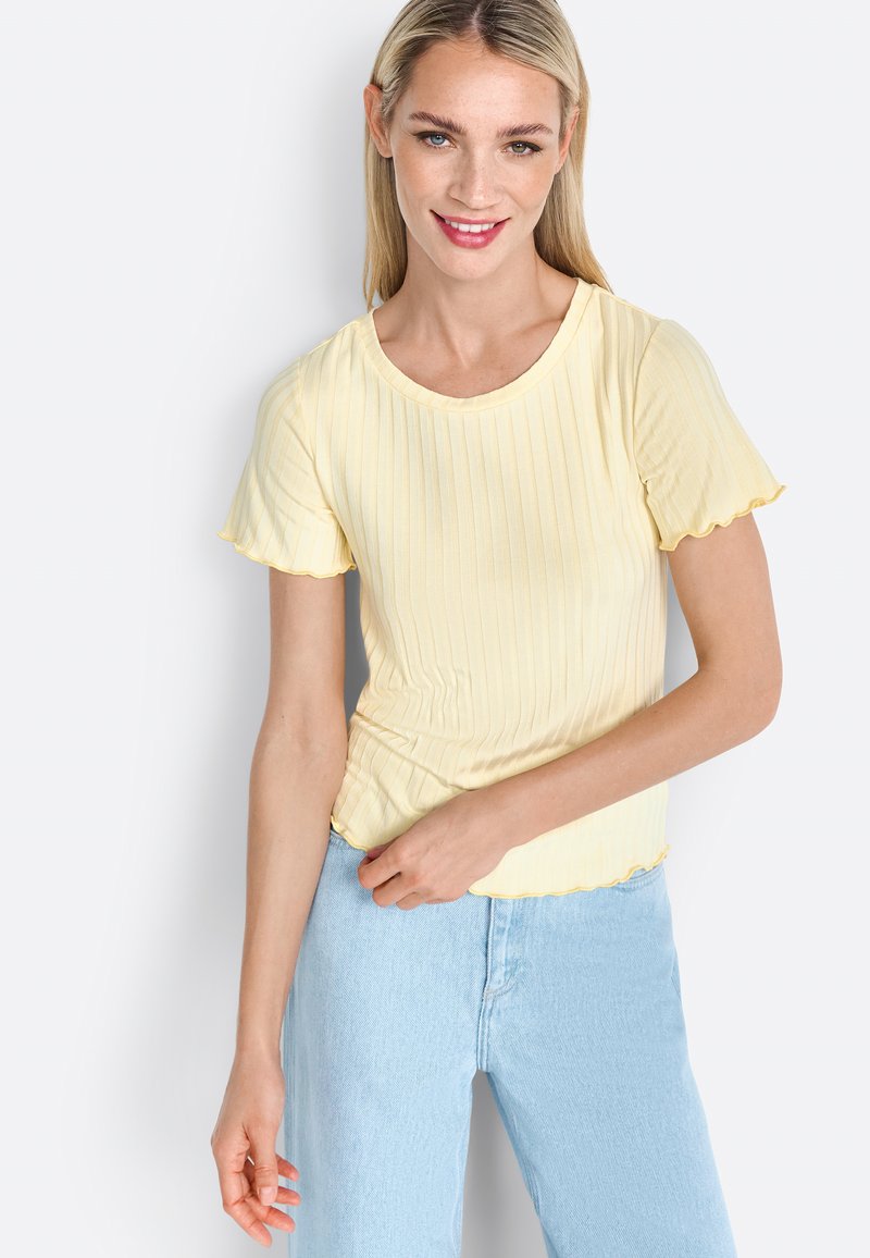 T-shirt giallo a righe con maniche corte e arruffate e scollatura tonda, abbinato a jeans azzurri. La T-shirt ha un orlo a onde.
