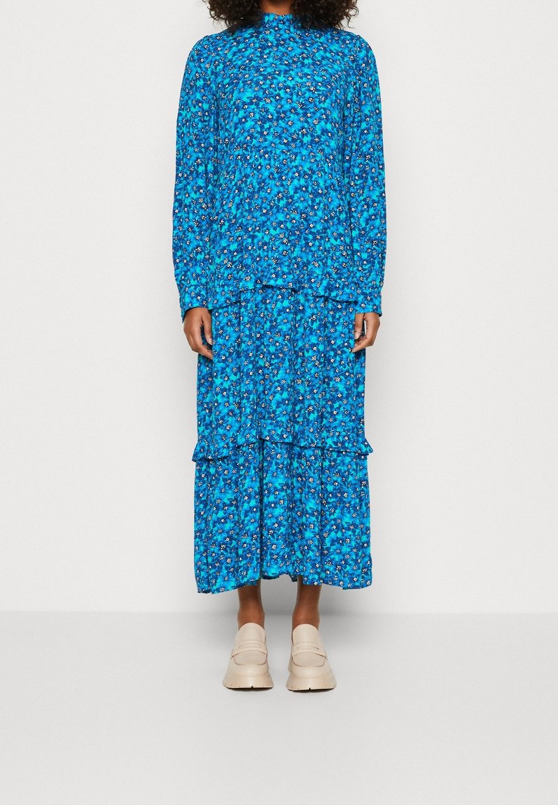 Femme portant une robe longue à manches longues bleu à fleurs avec des détails de volants, associée à des chaussures beige à enfiler, debout devant un fond uni.